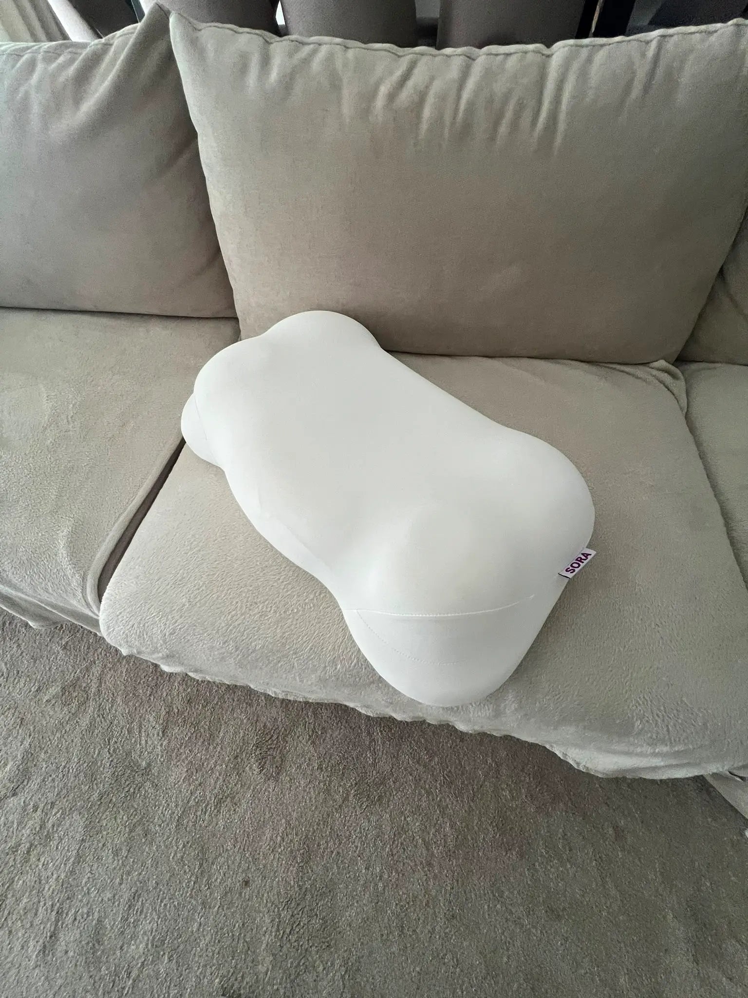 Real customer photo of SORA Cloud ergonomic pillow on a beige sofa — user-generated content showing bone-shaped memory foam pillow with SORA label in a home setting