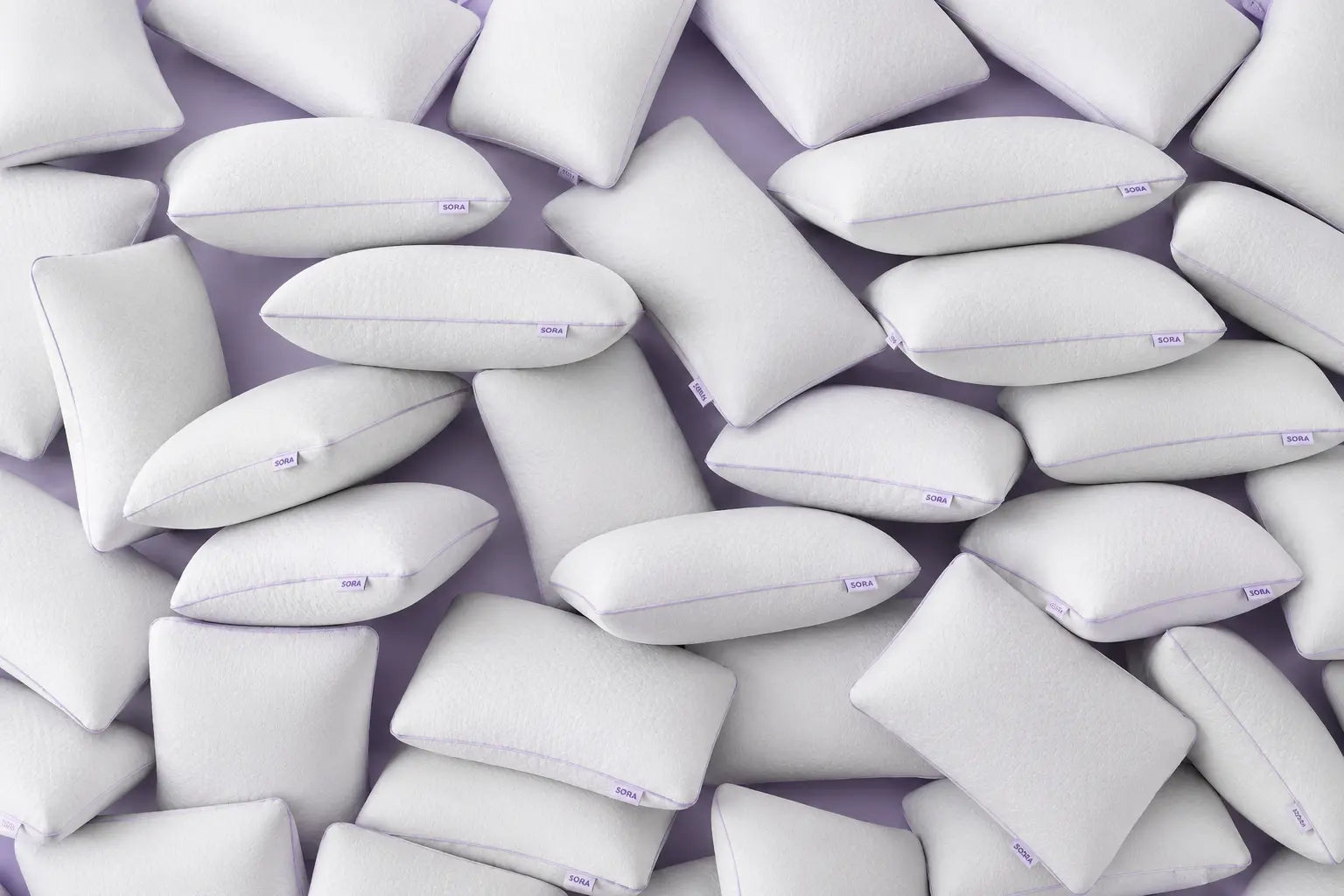 Multiple SORA branded pillows scattered on a lavender background — collection of white ergonomic sleep pillows with SORA labels