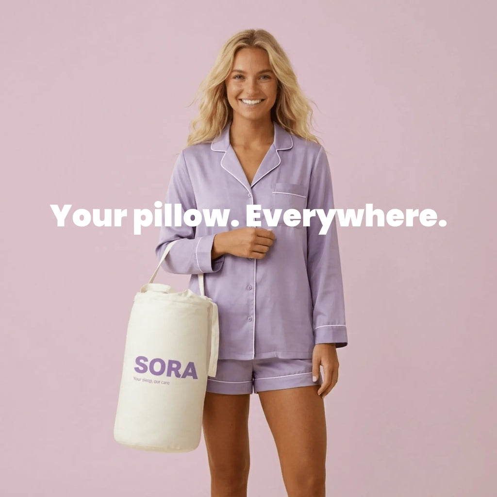 Smiling woman in lavender silk pijamas holding the SORA branded canvas travel bag by her side, standing against a soft pink background