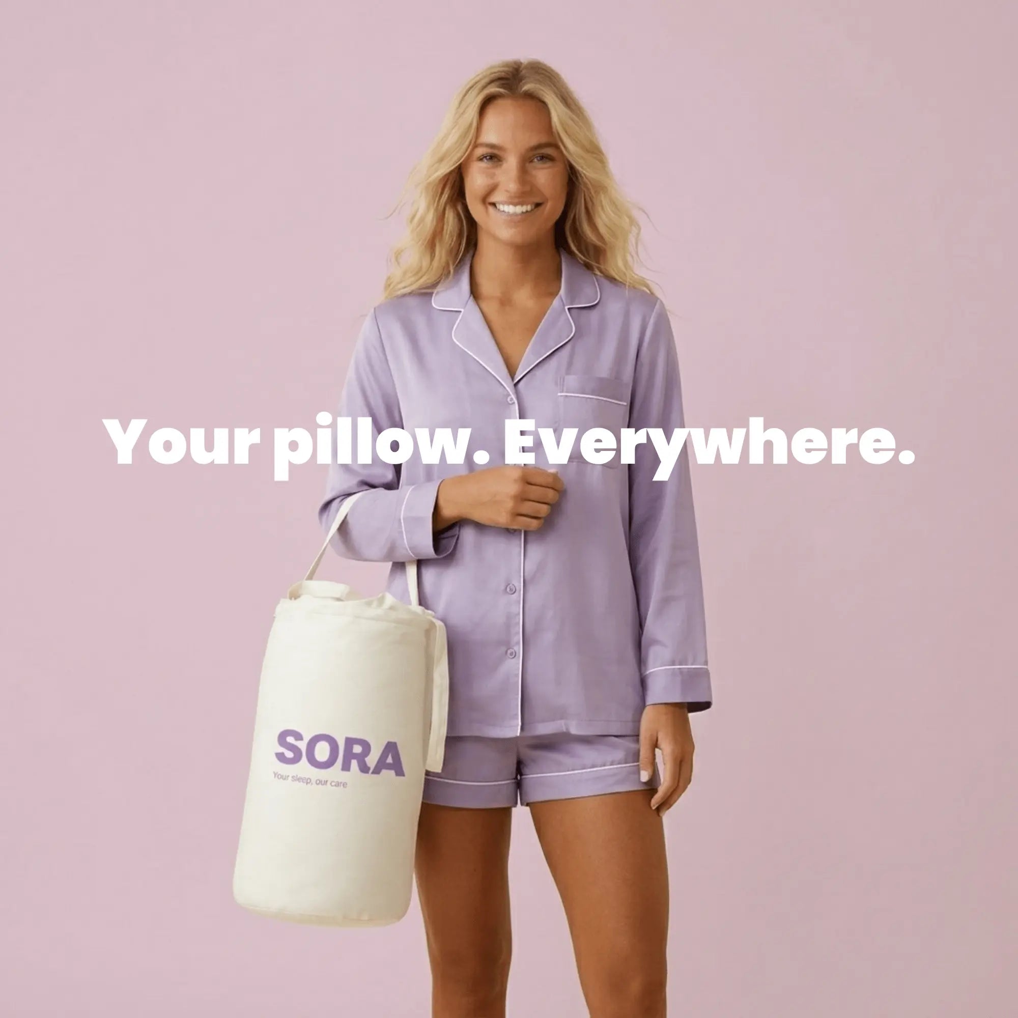 Smiling woman in lavender silk pijamas holding the SORA branded canvas travel bag by her side, standing against a soft pink background
