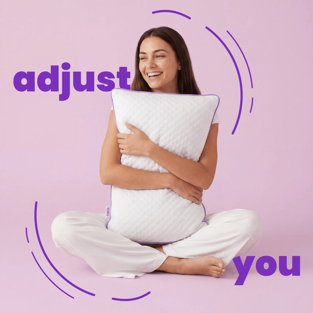 Woman sitting cross-legged hugging SORA Adjust ergonomic pillow on pink background